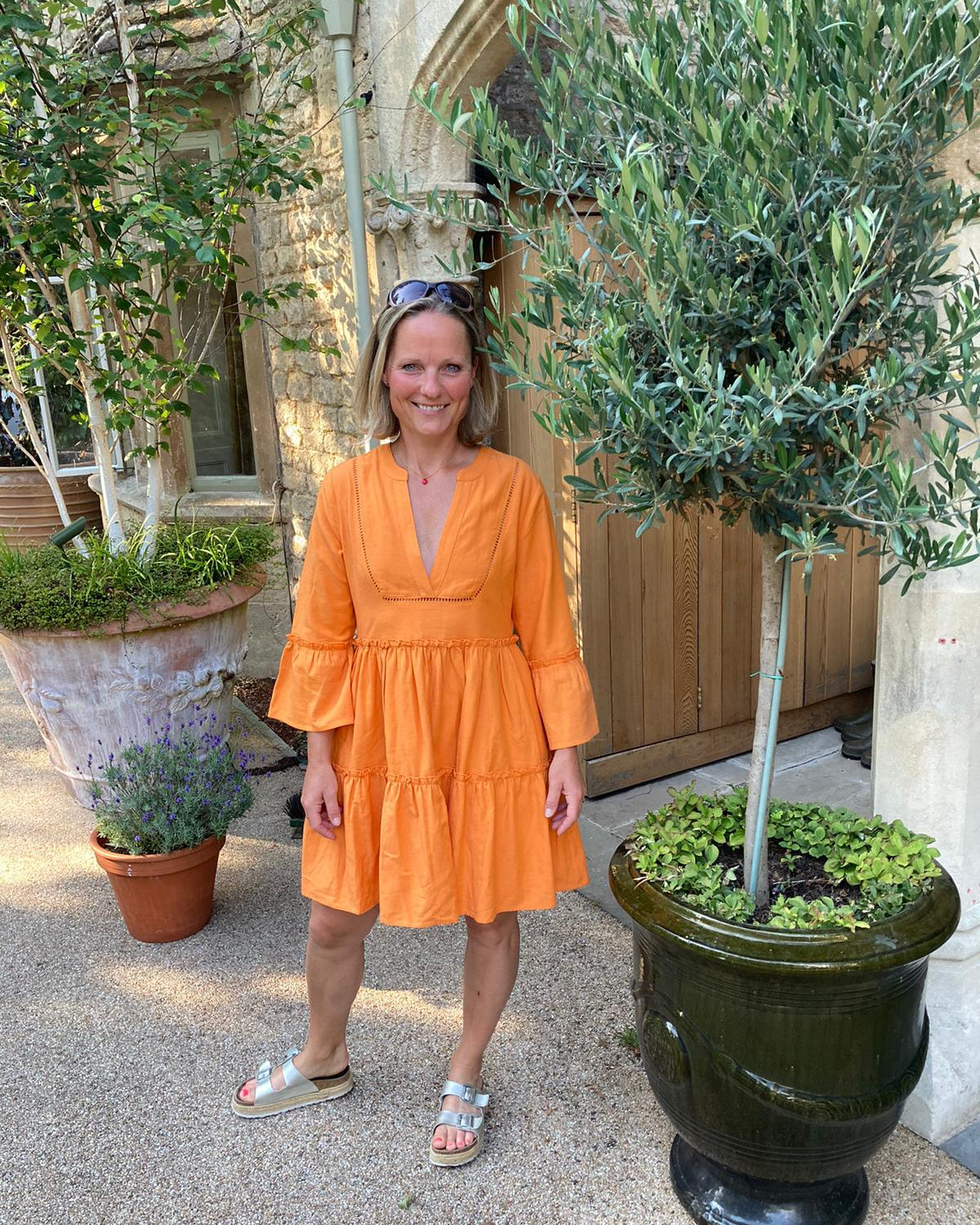 Woman in an orange dress standing outdoors with potted plants and a building in the background